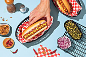 A close-up of hands holding a hot dog with mustard, surrounded by colourful toppings on a light blue background