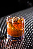 A classic whiskey Old Fashioned is served with a slice of dried citrus fruit in a clear glass The cocktail rests on a textured metal surface that emphasises its rich amber tones