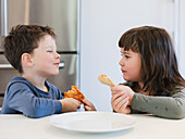 Two children eat happily at a kitchen table, holding slices of pizza in their hands. The scene captures their happiness and camaraderie in a bright kitchen environment.