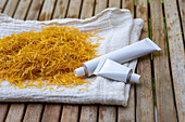 Marigold blossoms on a cloth and white cosmetic tubes with marigold blossom cream on a wooden surface. The picture shows a natural, plant-based approach to skin care and beauty products.