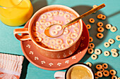Top view of a colourful breakfast service with a ceramic bowl of cereal and milk, a cup of coffee and orange juice on a turquoise surface.
