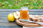 A rustic breakfast scene on a wooden table with a jar of quince jam, a knife, bread spread with jam and fresh quinces against a blurred natural green background.