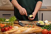 A person skilfully prepares an avocado, surrounded by fresh vegetables and bread on a kitchen counter. A delicious spread for a healthy meal is ready.