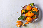 A bowl of fresh mandarins with green leaves, arranged on a textured surface. One mandarin is partially peeled, highlighting its bright orange colour and juicy texture.