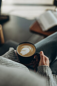 A person holds a latte with an artistic head of foam and relaxes in a cosy setting. The open book and soft jumper convey a sense of peace and leisure, perfect for coffee time