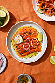 A beautifully presented salmon carpaccio with lime, black and white sesame seeds, cucumber and onion slices, which makes a lively presentation on an orange tablecloth.