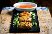 Close-up of golden corn and spring onion patties, served with fresh greens and a bowl of sweet chilli sauce The dish is elegantly presented on a slate board with a rustic background