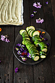 Fresh courgette slices and colourful edible flowers are arranged on a black plate, ready to garnish a cake on a rustic wooden table.