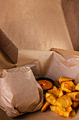 Close-up of a wrapped burger next to golden, crispy potato wedges and a small bowl of savoury dipping sauce, all on branded packaging. Perfect for a food advert.