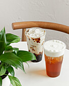 Two iced drinks, creamy coffee and frothy tea, are placed on a round white table next to a lush green plant A wooden chair emphasises the cosy, inviting atmosphere