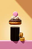 A visually striking stack of a chocolate donut with popcorn and a pink macaron placed over a glass of coffee Two macarons sit next to it, all against a vibrant background