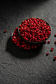 From above: A striking image showing a bowl of bright red peppercorns on a textured slate background. The dark tones contrast with the bright red of the peppercorns and emphasise their colour and texture.