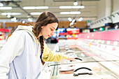 A woman is seen from the side as she examines the products in the frozen food section of a supermarket and apparently decides what she wants to buy.