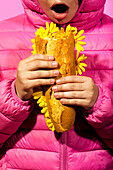 Ein auffälliges Bild eines Mädchens in einer rosafarbenen Jacke, das ein Baguette hält, das mit gelben Blumen vor einem rosafarbenen Hintergrund verwoben ist. Die leuchtenden Farben bilden einen optisch ansprechenden Kontrast.