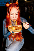 A woman with vibrant hair savours noodles with chopsticks at an Asian food market, capturing the essence of culinary discovery and cultural diversity