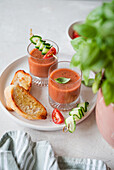 Two glasses of fresh gazpacho, garnished with cucumber and tomato skewers, served with toasted bread on a white plate A fresh basil leaf adds a splash of colour