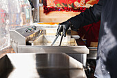 A hand in a black glove uses tongs to deep-fry spiral-shaped tornado potatoes in an outdoor snack bar. The stainless steel fryer and festive decorations create a lively market atmosphere.