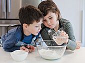 Two young siblings are baking while carefully measuring and mixing ingredients in a brightly lit kitchen. They wear aprons and concentrate on the task they are enjoying.
