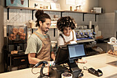 Two cheerful baristas in aprons work together at a café counter and operate a touchscreen cash register The cosy café ambience includes shelves, pastries and coffee accessories