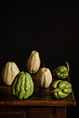 A rustic display of fresh chayote on a dark wooden table against a black background. The textures and colours of the vegetables convey a natural, organic aesthetic.