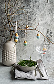 An Easter table setting with a ceramic vase containing painted eggs hanging from bare branches and a bowl of grass accentuated by the subtle texture of a tablecloth