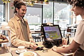 A café employee helps a smiling customer at the till. The atmosphere is inviting, with a display of pastries and a cosy café interior in the background