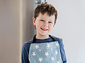 Ein fröhliches Kind trägt eine Schürze mit Sternenmuster und lächelt freundlich. Der Hintergrund ist leicht unscharf und unterstreicht den fröhlichen Gesichtsausdruck und die legere Kleidung der jungen Köchin.