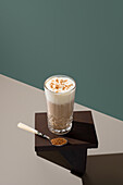 A crystal glass filled with creamy coffee, topped with milk cream and sprinkled with brown sugar, stands on a dark, angular surface A spoonful of sugar is included