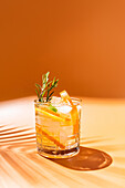 A lively summer cocktail with fresh citrus slices and a sprig of rosemary, presented in a glass against a backdrop of warm orange tones.