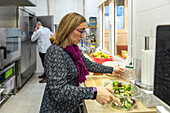 A woman prepares a fresh salad with a glass bowl in a modern kitchen. Her partner can be seen in the background, emphasising the lively, domestic atmosphere of a house under surveillance.
