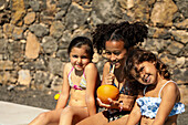 Drei Kinder in Badeanzügen sitzen fröhlich an einer Steinmauer. Sie genießen erfrischende Getränke, sonnen sich im Sonnenlicht und verkörpern die Freude am Sommerurlaub.