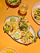 Draufsicht auf die Zubereitung einer Taco-Mahlzeit vor einem leuchtend gelben Hintergrund. Das Bild zeigt Tacos gefüllt mit Lachs, Mango, Koriander und Avocado