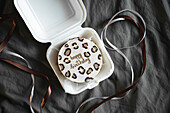 Top view of a stylish bento birthday cake with a happy birthday message, decorated with animal motifs. The cake is presented in a white takeaway box, complemented by elegant ribbons on a textured grey background.