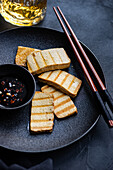 Grilled tofu slices on a black ceramic plate, served with a spicy dipping sauce and chopsticks, from the top. Perfect for presenting a vegan meal.