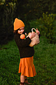 A small child wearing an orange cap and skirt carries a wicker basket filled with oranges. The scene is set in a lush, green environment and embodies the mood of autumn.