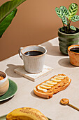 A quiet breakfast scene with a ceramic cup of coffee and a piece of toast topped with peanut butter and banana slices. A small plant adds a touch of nature.