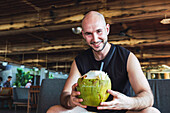 A cheerful bald man sips a fresh coconut while relaxing in a rustic tropical café that embodies the spirit of holiday and leisure in Bali.