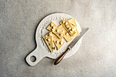 Top view of a white chocolate bar with hazelnuts, broken into pieces on a white vintage plate next to a rustic knife on a textured grey concrete background.