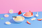 Vanilla and strawberry ice cream in white bowls surrounded by colourful paper flowers on a pink and blue background, accentuated with fresh strawberries and decorative elements.