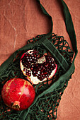 A bright red pomegranate stands on a green mesh bag against a background of brown fabric. The fresh fruit and rustic elements create a natural, organic impression.