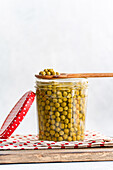 A jar filled with green, home-made organic peas, topped with a wooden spoon. The jar stands on a wooden tray with a red dotted cloth, creating a rustic kitchen scene.
