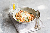 A ceramic bowl of cavatappi pasta topped with grated cheese, fresh basil leaves and spicy tomato sauce. On a grey surface, together with a glass of water and a slice of lemon.
