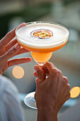 Eine Frau hält einen Passionsfruchtcocktail mit cremiger Schaumkrone in der Hand und fängt damit die Essenz eines luxuriösen Abends ein. Die verschwommene Hintergrundbeleuchtung schafft eine warme, intime Atmosphäre.