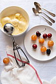 Top view of a refreshing dessert with a bowl of creamy vanilla ice cream, a plate of bright cherries, apricots and a blackberry lying on a white tablecloth next to old spoons.