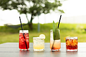 Eine Vielzahl bunter Cocktails in eleganten Gläsern steht auf einem Tisch im Freien vor einer natürlichen, verschwommenen grünen Kulisse - perfekt für Sommerstimmung und Entspannung.