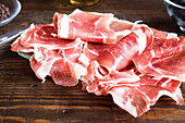 Thin, delicately rolled slices of fresh ham are presented on a dark wooden surface, highlighted by natural light that brings out the rich, pink tones and marbled texture of the meat.