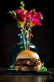 A gourmet pulled pork slider is elegantly presented with colourful flowers in the background. The food is garnished on a slate plate, creating an exquisite presentation.