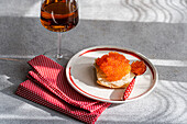 From above: a sumptuous presentation of red trout caviar on a slice of bread, accompanied by a glass of amber wine, on a dotted napkin and plate against a shaded background.