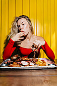 A woman in a bright red outfit enjoys a selection of grilled meat on a tray. The bright yellow background and her satisfied expression emphasise the joy of a barbecue.