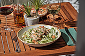 An appetising place setting with a Caesar salad with chicken and glasses of red and white wine, creating a perfect outdoor dining experience on a wooden table.